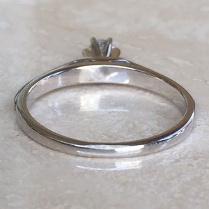 Pode incluir: Um anel de metal prateado com uma &uacute;nica pedra preciosa transparente e facetada, presa por uma crava&ccedil;&atilde;o de m&uacute;ltiplas garras. A banda &eacute; lisa e polida, exibindo uma superf&iacute;cie reflexiva que capta a luz com a pedra na parte superior.