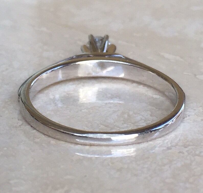 Pode incluir: Um anel de metal prateado com uma &uacute;nica pedra preciosa transparente e facetada, presa por uma crava&ccedil;&atilde;o de m&uacute;ltiplas garras. A banda &eacute; lisa e polida, exibindo uma superf&iacute;cie reflexiva que capta a luz com a pedra na parte superior.