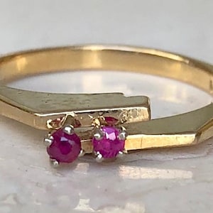 May include: A gold bypass ring featuring two small, round, faceted red rubies set in silver-toned prongs. The band has a smooth, polished finish with a distinct angular design where the two ends overlap, showcasing a clean and simple construction.