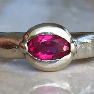 May include: A polished silver-toned ring featuring a central oval-cut vibrant pink gemstone set in a smooth bezel setting. The band is solid and rounded, showcasing a reflective metallic finish that highlights the bright color of the stone.