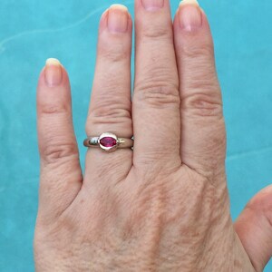 May include: A silver-toned ring featuring a single, horizontally set, oval-shaped, vibrant pinkish-red gemstone. The stone is secured within a smooth, polished metal bezel setting that sits flush against the band of the ring.