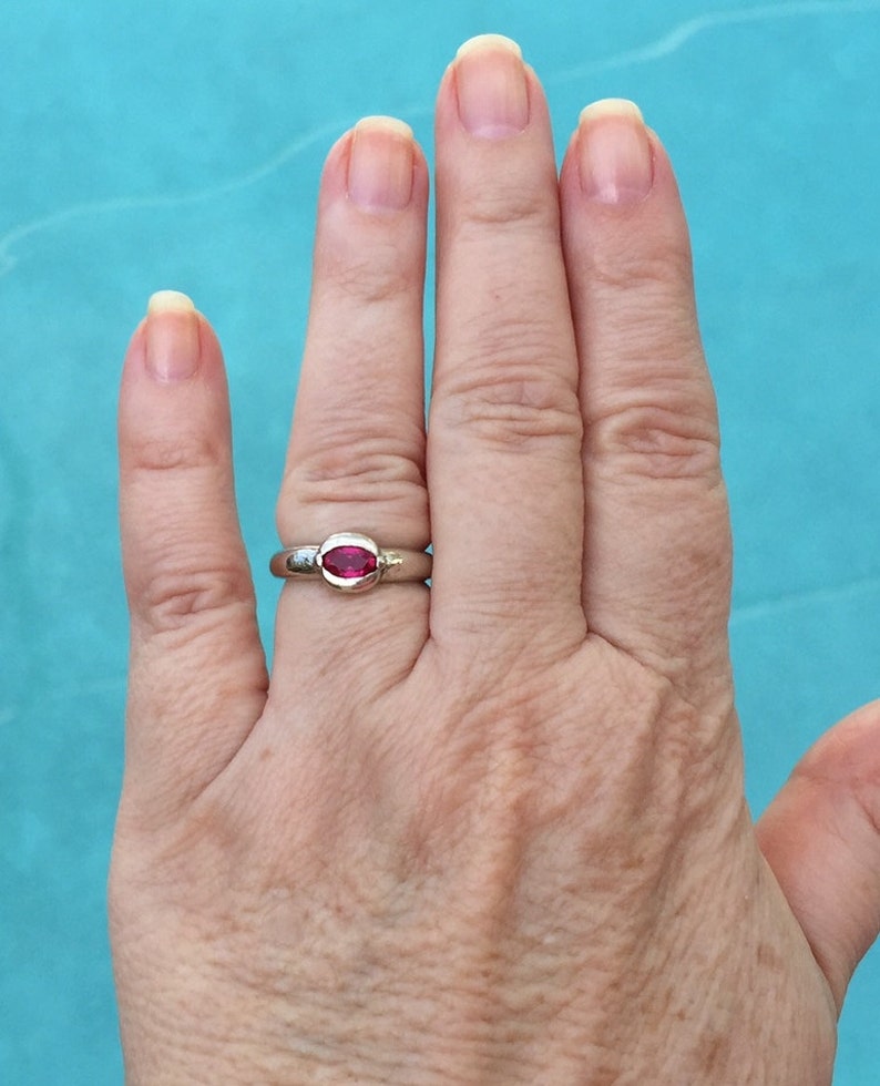 May include: A silver-toned ring featuring a single, horizontally set, oval-shaped, vibrant pinkish-red gemstone. The stone is secured within a smooth, polished metal bezel setting that sits flush against the band of the ring.