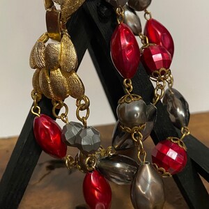 May include: A vintage bracelet featuring gold-tone metal leaf accents and multiple strands of beads. The beads are in shades of red, gray, and silver, with varying shapes and textures. The bracelet is displayed on a black stand.