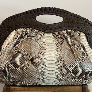 May include: A brown and white snakeskin clutch purse with a brown leather top handle. The purse has a textured snakeskin pattern and a unique, slightly rounded shape.