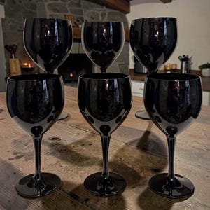 May include: A set of five matching wine glasses made of solid black glass. Each glass features a smooth, rounded bowl, a slender stem, and a circular base. The glasses have a glossy, reflective surface that catches light, creating a uniform appearance.