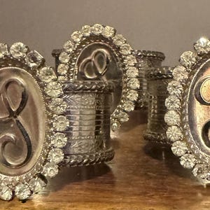 May include: A set of silver-toned metal napkin rings, each featuring an oval centerpiece with a prominent engraved letter S. The oval plaque is surrounded by a border of clear, faceted rhinestones, and the band has a textured, etched finish.