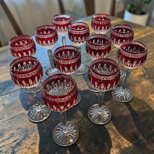 May include: A set of eleven crystal wine glasses featuring a deep red overlay on the bowl with intricate cut glass patterns, including vertical flutes and diamond motifs, supported by clear glass stems and circular bases with a starburst design.