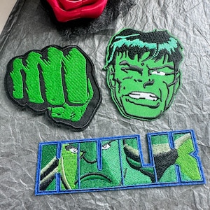 Könnte beinhalten: Drei gestickte Aufnäher mit dem Hulk-Motiv. Ein Aufnäher zeigt eine grüne geballte Faust, ein weiterer das Gesicht der Figur mit schwarzen Haaren und grüner Haut, und der dritte ist ein rechteckiger Aufnäher mit dem Wort HULK in blauen Rändern.