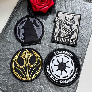 Puede incluir: Cuatro parches de tela bordados con temas de Star Wars. Un parche circular muestra una Estrella de la Muerte negra y gris, un parche rectangular representa un soldado de asalto blanco con el texto TROOPER, un símbolo circular dorado y negro de la Alianza Rebelde y un parche circular negro y blanco de Republic Commando.