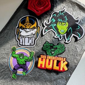 Puede incluir: Cuatro parches bordados con personajes de cómic. Uno muestra a un villano de piel morada, otro a un personaje de piel verde con pelo negro, otro a una figura musculosa verde en un círculo y otro con el texto The Incredible Hulk.