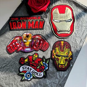 Puede incluir: Cinco parches bordados con imágenes de Iron Man. Un parche rectangular dice THE INVINCIBLE IRON MAN. Otros incluyen una máscara de casco, una figura voladora, una máscara estilizada con patrones florales y una figura con el texto STAY GOLD.