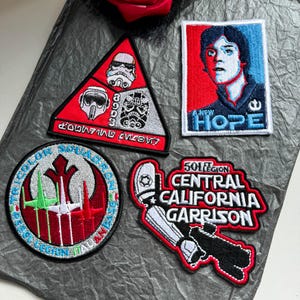 Puede incluir: Cuatro parches bordados con temas de Star Wars. Un parche triangular muestra tres cascos. Un parche rectangular representa a un personaje con el texto A NEW HOPE. Un parche circular muestra tres cazas estelares. Un parche con forma lee 501ST LEGION CENTRAL CALIFORNIA GARRISON.