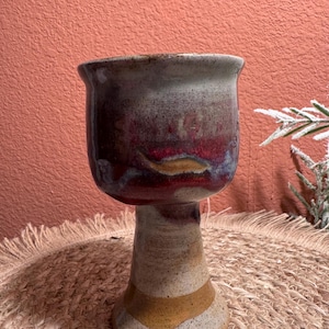 May include: A ceramic goblet featuring a speckled off-white base with a wide, flared rim. The upper bowl displays a blended glaze of deep crimson, dark purple, and earthy brown tones, while the stem and circular foot feature horizontal bands of ochre.