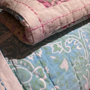 Könnte beinhalten: Ein gefaltetes Stepptextil mit einem rosa-grünen Blumenmuster und einem cremefarbenen Rand, der mit rosa Faden bestickt ist. Darunter liegt ein zweites Stepptextil mit einem hellblauen und weißen Blumenmuster und blauen Nähten.