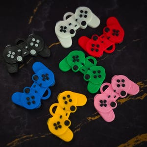May include: A collection of seven small, flat, plastic video game controller charms in various colors including black, white, red, green, blue, yellow, and pink. Each charm features a loop at the top for attachment and molded button details.
