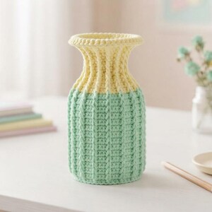 May include: A cylindrical vase featuring a textured crochet exterior with a ribbed pattern. The upper section is pale yellow, while the lower portion is a soft mint green. The vase has a flared rim and a straight body, crafted from yarn.