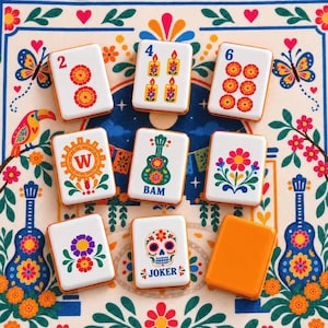 May include: A set of rectangular mahjong tiles featuring colorful illustrations of flowers, candles, a guitar, a skull, and the letter W. The tiles are white with orange edges and are arranged on a patterned fabric surface with floral and bird motifs.