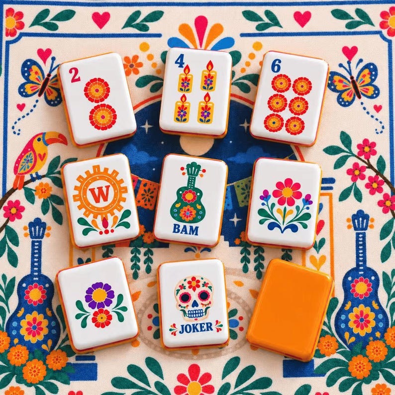 May include: A set of rectangular mahjong tiles featuring colorful illustrations of flowers, candles, a guitar, a skull, and the letter W. The tiles are white with orange edges and are arranged on a patterned fabric surface with floral and bird motifs.