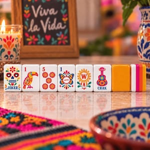 May include: A row of seven rectangular game tiles featuring colorful folk art designs, including a sugar skull labeled JOKER, a toucan, a number five, a butterfly, a letter W, a guitar labeled CRAK, and two solid orange and striped tiles.
