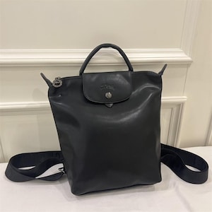 May include: A black leather backpack featuring a top handle, a fold-over flap with a silver-toned snap button closure, and adjustable shoulder straps. The bag includes a zippered main compartment with a circular silver-toned zipper pull.