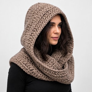 May include: A light brown hooded cowl featuring a thick, textured crochet stitch pattern. The garment is designed to drape around the neck and shoulders, with a deep hood that frames the head, crafted from a solid, neutral-toned yarn.