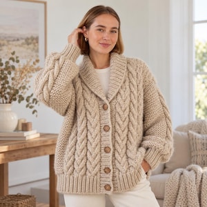Peut inclure: Un cardigan en tricot beige épais doté d'un col châle, de manches longues à poignets côtelés et d'un motif torsadé vertical. L'avant est fermé par cinq gros boutons ronds marron, et l'ourlet est fini par une large bande en tricot côtelé.