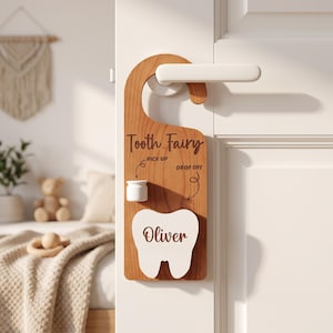 May include: A wooden door hanger featuring the text Tooth Fairy, Pick Up, and Drop Off. It includes a small white container and a white tooth-shaped cutout with the name Oliver engraved in brown. The hanger is designed to hook over a door handle.