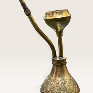 May include: A polished brass smoking pipe featuring a wide, funnel-shaped bowl atop a bulbous base. The base is decorated with intricate embossed floral and geometric patterns. A long, slender, curved stem extends upward with a textured grip area.