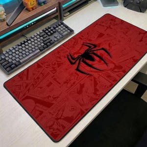 May include: A rectangular desk mat featuring a monochromatic red comic book panel print with a large black spider emblem centred on the surface. The mat is positioned on a desk next to a black mechanical keyboard and a small toy figure.