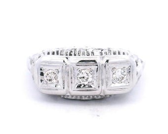 May include: A white gold ring featuring three square-cut diamonds set in a row. The ring has an ornate, filigree design on the band and around the diamond settings. The diamonds sparkle against the white metal.