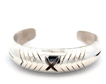 May include: A silver cuff bracelet with an engraved geometric pattern and an X-shaped design in the center. The bracelet has a rounded shape and is open at the ends.