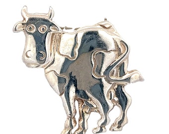 May include: A silver cow brooch with a detailed design. The cow is standing with its head turned to the side and its tail curled up.