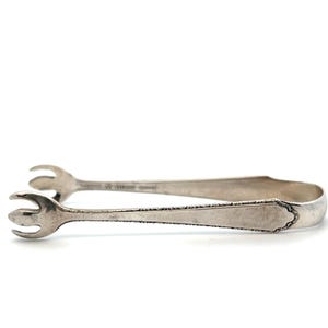 May include: Silver-toned sugar tongs with a decorative edge and claw-like ends. The tongs are designed to grip and lift sugar cubes. The design is simple and elegant, suitable for serving sugar at a tea party or coffee gathering.