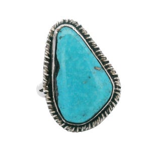 May include: A sterling silver ring featuring a large, irregular, teardrop-shaped turquoise stone with a vibrant blue color. The stone is set within a bezel and surrounded by a decorative twisted rope-style metal border, creating a textured frame.