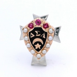 May include: A gold and silver pin featuring a black shield with white Greek letters, a crescent moon, and a star. The pin is adorned with pink gemstones and surrounded by pearls. The pin has a silver bow-like design on the top and bottom.