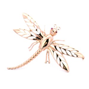 May include: A gold-toned dragonfly brooch with intricate detailing. The dragonfly features openwork wings with white enamel, a segmented body, and small red accents for the eyes. The brooch is set against a white background.