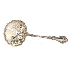 May include: A silver serving spoon with an ornate, pierced bowl and a long, detailed handle. The bowl has a decorative, openwork design. The handle features intricate patterns and a rounded end. The spoon is likely for serving sauces or other foods.