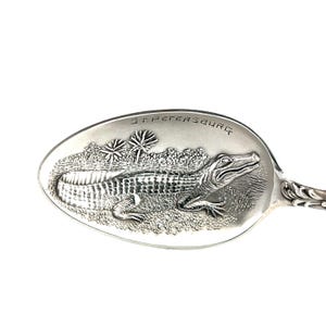 May include: Silver spoon with an embossed design featuring an alligator and palm trees. The text "ST. PETERSBURG" is engraved above the alligator. The spoon has a decorative handle.