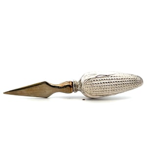 May include: A silver-toned metal letter opener featuring a handle shaped like a textured ear of corn. The blade is pointed and tapers to a sharp tip, while the handle displays detailed kernels and husks in a metallic finish with slight patina.