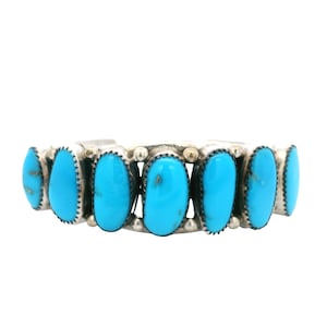 May include: A silver cuff bracelet featuring seven oval turquoise stones. The bright blue stones are set in individual bezels and are surrounded by a decorative silver border. The bracelet is a classic piece of Southwestern jewelry.