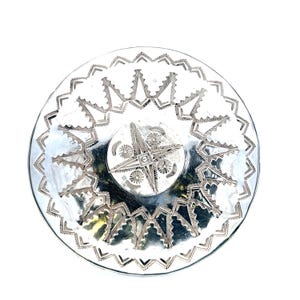 May include: A circular silver-toned metal concho featuring a central four-pointed star design surrounded by small circular motifs. The outer edge is decorated with a repeating geometric zigzag pattern, creating a textured, reflective surface.
