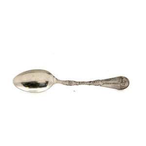 May include: A silver spoon with an oval bowl and a detailed handle. The handle features intricate patterns and a small emblem at the end. The spoon is likely vintage or antique, with a classic design.