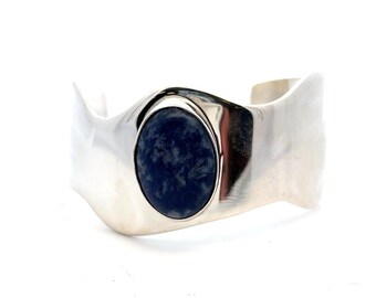May include: A wide, polished silver cuff bracelet featuring an oval, dark blue stone. The bracelet has a smooth, flowing design with a central setting for the stone. The cuff is open at the back.