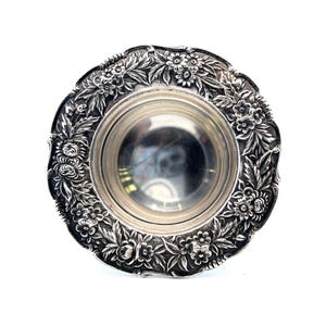 May include: A silver bowl with a floral design around the rim. The bowl is round and has a smooth, reflective surface.