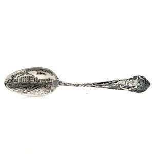 May include: A silver souvenir spoon with an engraved scene of a building and the text "Sault Ste. Marie, Mich." on the bowl. The handle features a detailed design of a Native American figure.