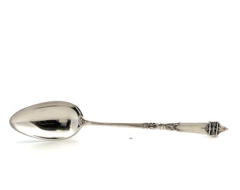 May include: A silver spoon with an oval bowl and a long, slender handle. The handle features ornate detailing and a rectangular end with a decorative design. The spoon is likely made of sterling silver.