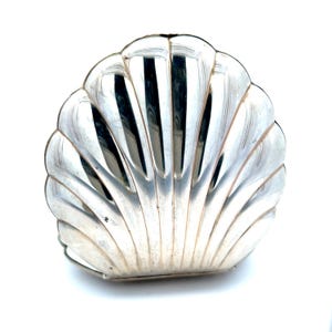 May include: A silver-colored, shell-shaped dish with a reflective surface. The dish has a scalloped edge and a detailed, ribbed design, creating a textured appearance. The dish is likely made of metal and sits on a white background.