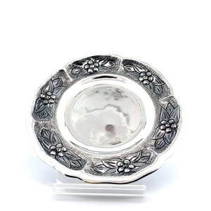 May include: A small, round, silver-colored dish with a scalloped edge. The rim is decorated with a repeating pattern of flowers and leaves. The dish is displayed on a clear stand.