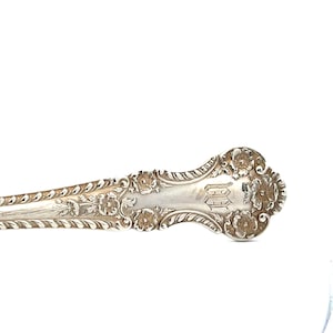 May include: Close-up of a silver spoon with ornate floral detailing. The spoon features a decorative handle with embossed flowers and a monogram. The spoon is made of a shiny, reflective metal.