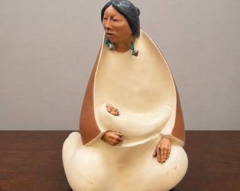 Puede incluir: Una escultura de cerámica de una figura sentada con cabello oscuro, que lleva pendientes de color turquesa y plateado, envuelta en un chal color crema con el exterior marrón. La figura sostiene a un pequeño bebé contra el pecho.